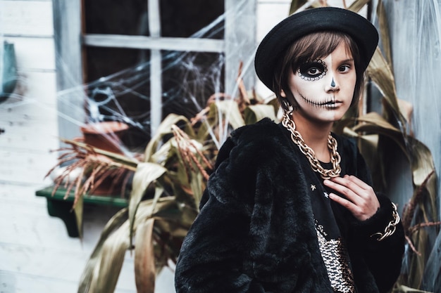 Bella bambina spaventosa che celebra halloween Terrificante trucco a metà viso bianco nero e costume da strega immagine elegante Divertimento horror alla festa per bambini nel fienile sulla strada Cappello catena di pelliccia