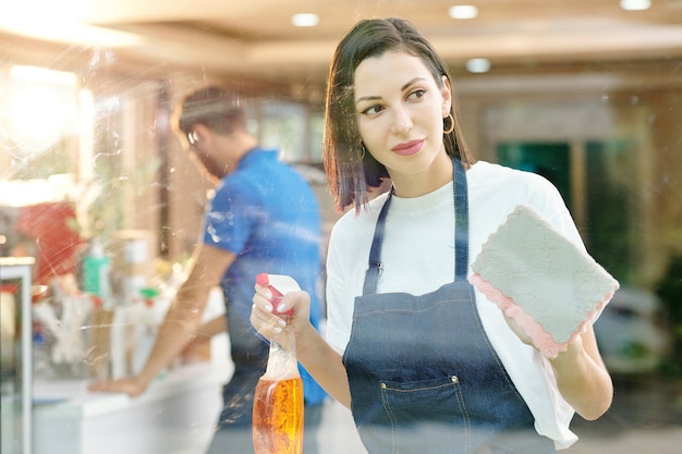 Bella barista femminile che spruzza detersivo sulla parete di vetro e lo pulisce con una spugna