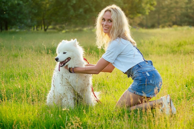 Bella bionda riccia sorridente felice giovane donna in pantaloncini di jeans formazione e giocando un cane samoiedo carino lanuginoso bianco sullo sfondo del campo di raggi tramonto del parco estivo. animale domestico e hostess.