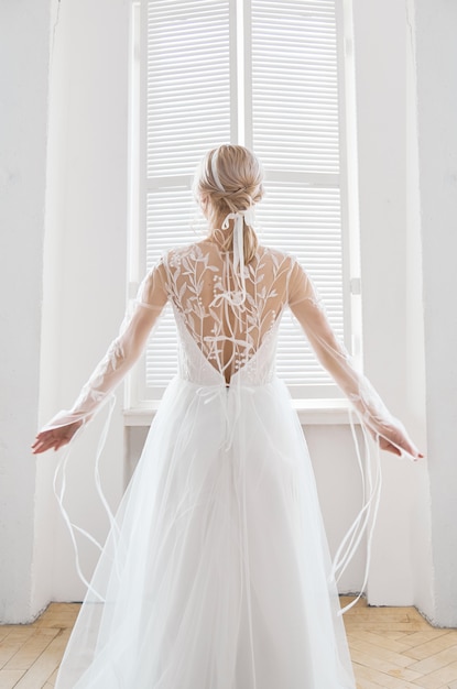 Bella bionda snella sotto il sole della sera in un lungo abito bianco vicino a una grande finestra. Capelli e trucco perfetti per la sposa, una nuova collezione di abiti da sposa