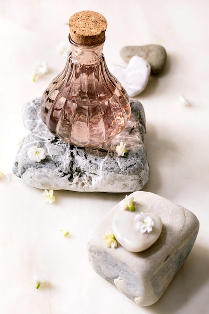 Bella bottiglia di profumo rosa trasparente con tappo in sughero in piedi su pietra strutturata su tavolo in marmo bianco con piccoli fiori bianchi Kalanchoe intorno al concetto di creazione di profumi con posto per etichetta di design