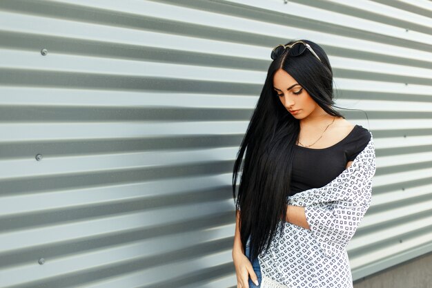 Bella bruna con i capelli lunghi in abiti alla moda vicino a un muro di metallo in strada