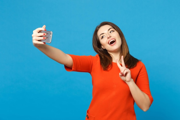 Bella bruna giovane donna che indossa un abito rosso arancione facendo selfie girato sul telefono cellulare isolato su sfondo blu alla moda, ritratto in studio. Concetto di moda stile di vita delle persone. Mock up spazio di copia.