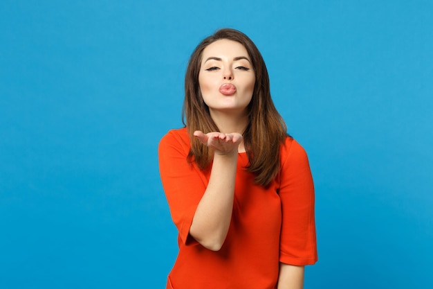 Bella bruna giovane donna che indossa un abito rosso arancione guardando la fotocamera che soffia l'invio di bacio d'aria isolato su sfondo blu parete ritratto in studio. Concetto di moda stile di vita delle persone. Mock up spazio di copia