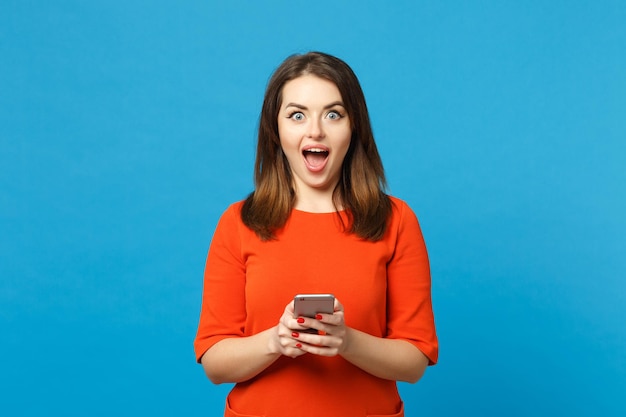 Bella bruna giovane donna che indossa un vestito rosso arancione messaggistica utilizzando il telefono cellulare isolato su sfondo blu alla moda della parete, ritratto in studio. Concetto di moda stile di vita delle persone. Mock up spazio di copia.
