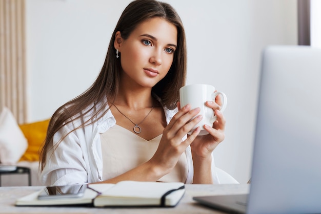 Bella bruna in camicia bianca si siede sul divano bianco, tenendo la tazza bianca con caffè caldo in mano.