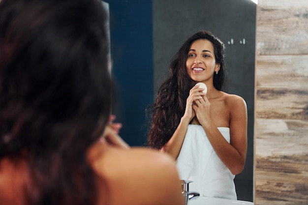 Bella bruna in piedi in bagno vicino allo specchio e pulendo il viso.