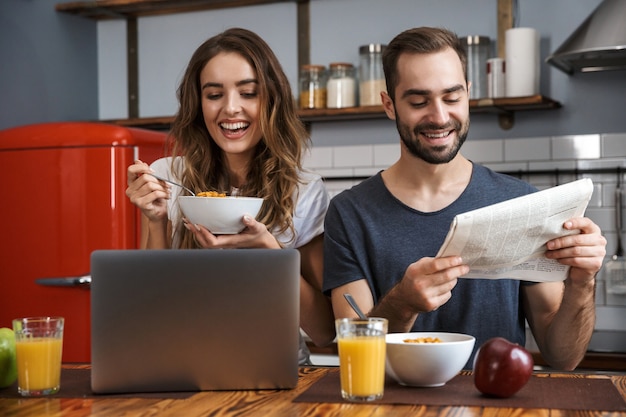 Bella coppia allegra facendo colazione in cucina, utilizzando il computer portatile, leggendo il giornale