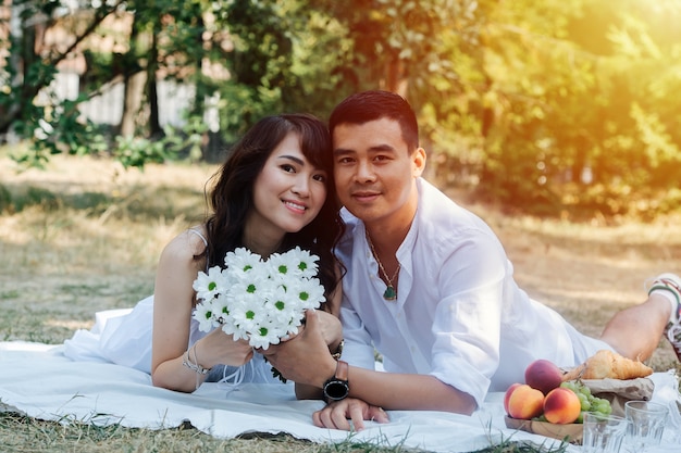 Bella coppia asiatica che fa un picnic in un parco, sdraiata a pancia in giù. Donna che tiene il bouquet, che riposa in abiti bianchi all'ombra di un albero.