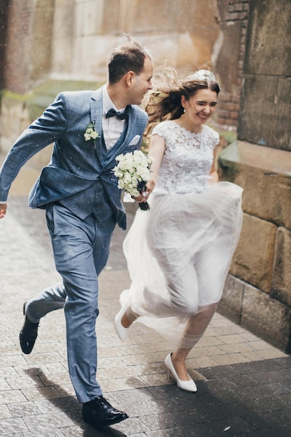 Bella coppia di sposi emotivi che sorridono e si divertono sotto la pioggia nella città europea Matrimonio in Provenza Elegante sposa e sposo felici che corrono sullo sfondo della vecchia chiesa in strada piovosa