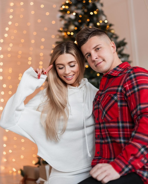 Bella coppia felice in abiti alla moda per la casa con camicia e felpa con cappuccio davanti alle luci di Natale e all'albero di Natale