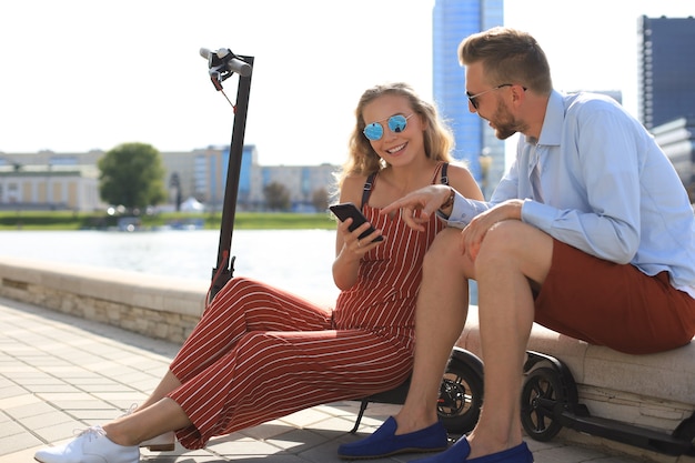 Bella coppia in vacanza divertendosi alla guida di uno scooter elettrico, prendendo una pausa dalla guida, seduta sulla riva del fiume.