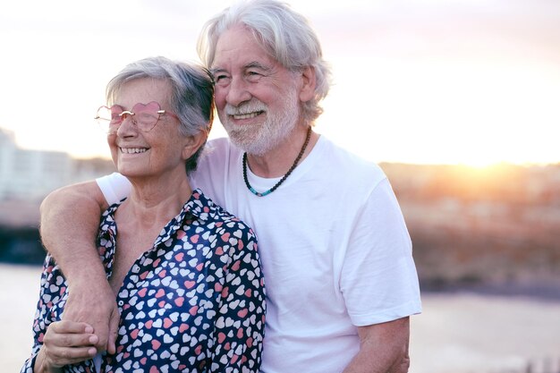 Bella coppia senior caucasica sorridente all'aperto in mare che guarda lontano. Coppia di anziani dai capelli bianchi che si gode la pensione e la luce del tramonto