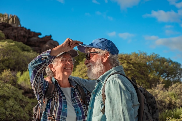 Bella coppia senior felice che abbraccia nel trekking in montagna godendosi la libertà della natura e uno stile di vita sano Vecchi pensionati sorridenti in cappello e abiti casual tra cespugli verdi e cielo blu