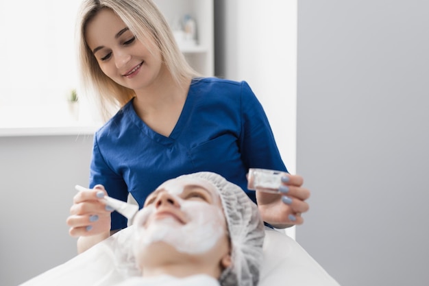 Bella cosmetologa femminile che applica una maschera crema idratante bianca sul viso con un pennello a un paziente sorridente