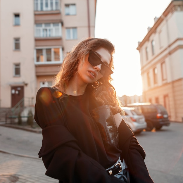 Bella donna abbastanza giovane hipster in abiti alla moda in stile retrò in occhiali da sole scuri alla moda in città in una giornata di sole.