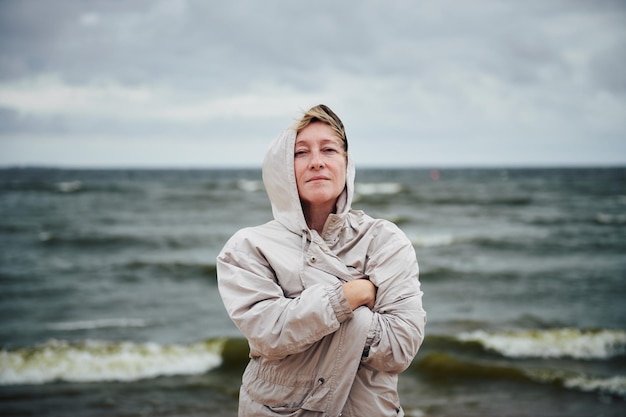 Bella donna adulta con un cappuccio e avvolta in una giacca sullo sfondo di un mare in tempesta
