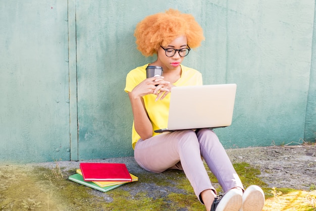 Bella donna africana con i capelli ricci che studia con laptop e libri seduti all'aperto sullo sfondo del muro verde