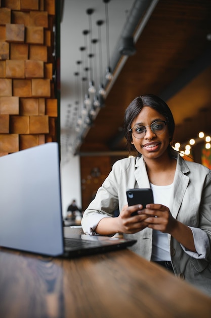 Bella donna africana con lunghi capelli scuri sta digitando messaggi su uno smartphone mentre è seduta accanto alla finestra in una luce soffusa Giovane donna d'affari sta usando laptop e telefono cellulare per il lavoro