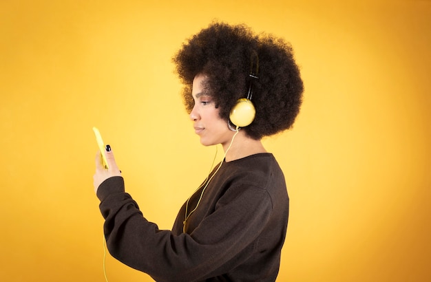 Bella donna afro che ascolta musica con cuffie e smartphone, sfondo giallo