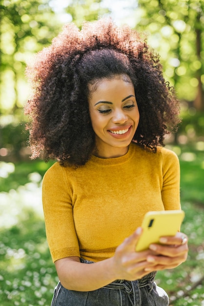 Bella donna afro digitando sul cellulare, nella foresta. Messa a fuoco selettiva.