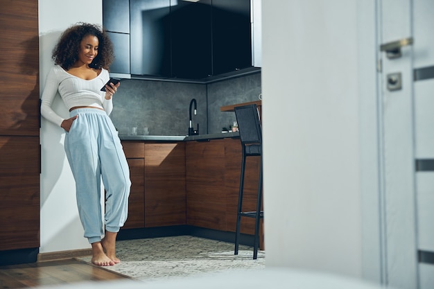 Bella donna afroamericana appoggiata al muro in cucina mentre tiene il cellulare e sorride