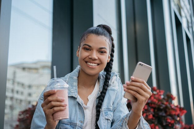 Bella donna afroamericana che tiene vetro con frullato e smartphone