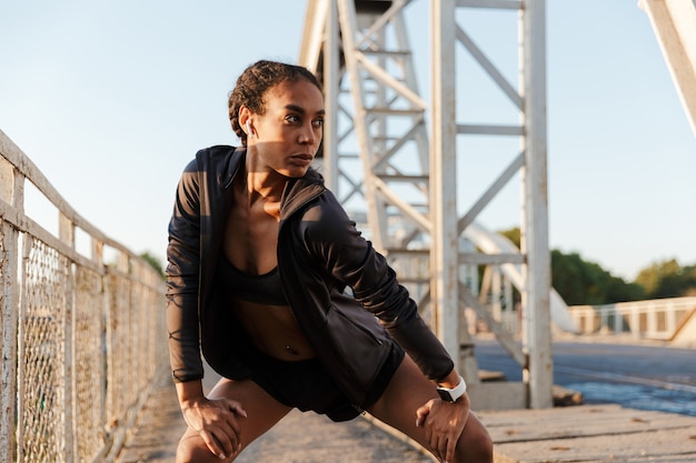 bella donna afroamericana in abbigliamento sportivo che utilizza auricolari e fa esercizi mentre si allena sul vecchio ponte