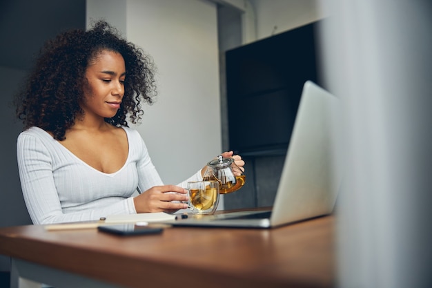 Bella donna afroamericana seduta vicino al suo computer portatile e sorridente mentre versa il tè nella tazza