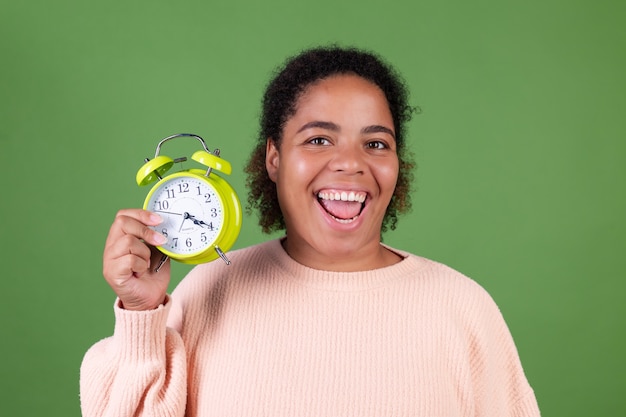 Bella donna afroamericana sulla parete verde con la sveglia sorridendo felice allegro