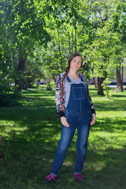 Bella donna all'inizio della gravidanza in tuta di jeans in piedi sull'erba verde nel parco