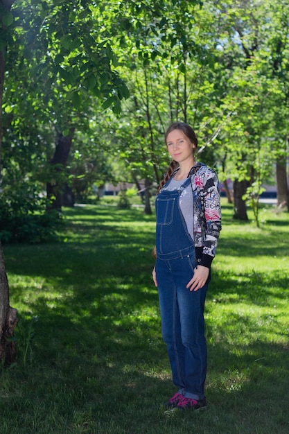Bella donna all'inizio della gravidanza in tuta di jeans in piedi sull'erba verde nel parco