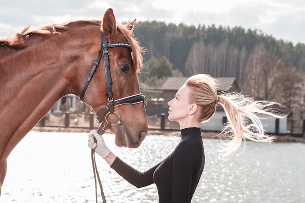 Bella donna alla moda che cammina con un cavallo in un country club. Sport equestri, noleggio di cavalli, concetto di tempo libero. Tecnica mista