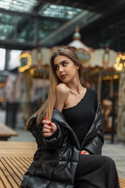 Bella donna alla moda in un top monospalla nero sexy alla moda con un piumino si siede su una panchina vicino a una giostra vintage con luci in città