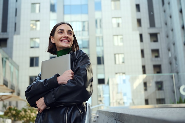 Bella donna allegra di successo, uomo d'affari, libero professionista, imprenditore, copywriter che tiene il laptop in mano e sorride graziosamente guardando da parte sullo sfondo degli edifici aziendali dell'ufficio moderno