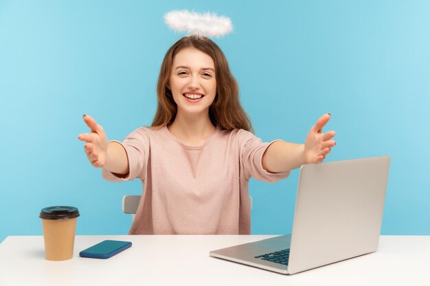 Bella donna angelica dipendente con nimbo seduto sul posto di lavoro e mani tese da abbracciare, abbracci liberi, segretaria che dà il benvenuto alla reception. tiro in studio indoor isolato su sfondo blu