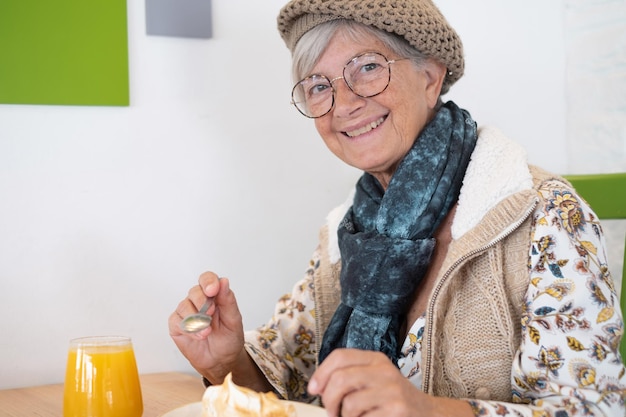 Bella donna anziana caucasica che si siede per la colazione nella femmina sorridente del caffè che mangia una torta al limone con un succo d'arancia