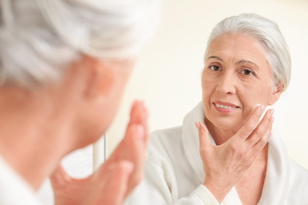 Bella donna anziana che applica crema per il viso davanti allo specchio a casa