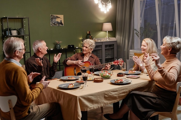 Bella donna anziana che suona la chitarra acustica mentre si siede al tavolo da pranzo nel soggiorno decorato e cena festiva con i suoi quattro amici