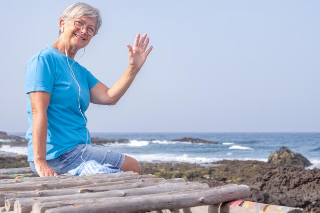 Bella donna anziana con gli occhiali seduto sulla spiaggia ascoltando musica con gli auricolari anziana donna sorridente guardando il concetto di pensionamento della fotocamera