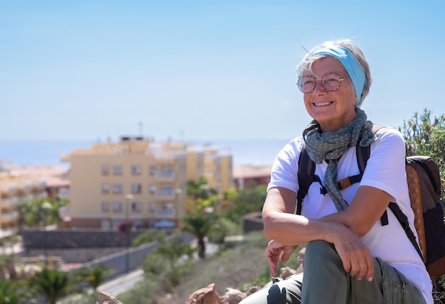 Bella donna anziana felice in un'escursione all'aperto che indossa uno zaino seduto con le braccia incrociate Orizzonte sul mare sullo sfondo