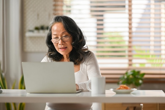 Bella donna anziana imprenditrice che lavora a distanza da casa leggendo la posta elettronica sul computer portatile