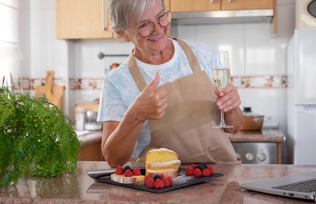 Bella donna anziana nella cucina di casa dopo aver cotto il suo plumcake fatto in casa con in mano un bicchiere di vino mentre chatta con gli amici o la famiglia sul computer Cucinare a casa concetto