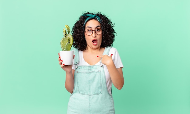Bella donna araba che sembra scioccata e sorpresa con la bocca spalancata, che indica se stessa e tiene in mano un cactus in vaso