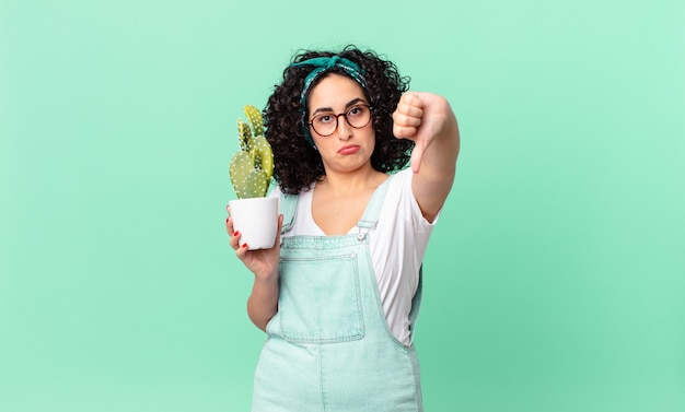 Bella donna araba che si sente arrabbiata, mostra i pollici in giù e tiene in mano un cactus in vaso