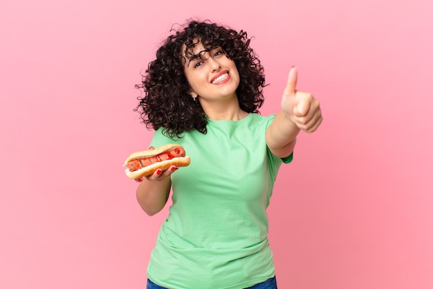 Bella donna araba che si sente orgogliosa, sorride positivamente con il pollice in alto e tiene in mano un hot dog