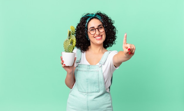 Bella donna araba che sorride e sembra amichevole, mostra il numero uno e tiene in mano un cactus in vaso