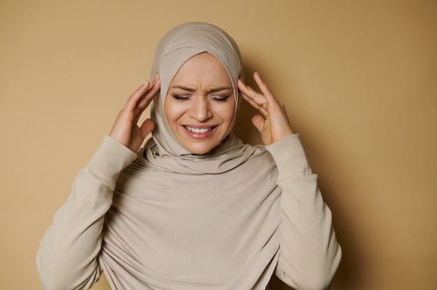 Bella donna araba in hijab in posa davanti esprimendo dolore e mal di testa su una superficie beige con spazio di copia