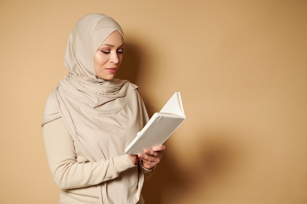 Bella donna araba musulmana che indossa l'hijab e vestito tradizionale formale rigoroso leggendo un libro sulla superficie beige con lo spazio della copia