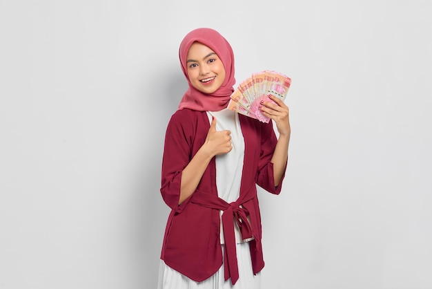 Bella donna asiatica allegra in camicia casual e hijab che tiene banconote in rupia indonesiana, mostrando il pollice in alto gesto isolato su sfondo bianco. Concetto di stile di vita religioso della gente